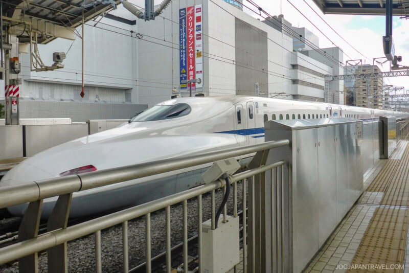 A sleek white Japanese bullet train (Shinkansen) with a blue stripe speeds past a platform at an urban station, as travelers clutching Japan train tickets admire the modern buildings in the background.