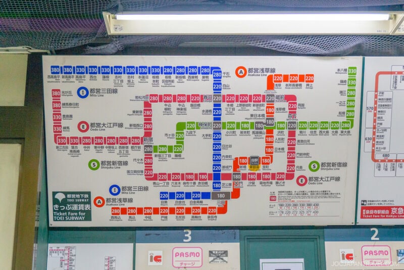 A Japanese subway fare map shows station names, colored subway lines, and fare prices in yen—perfect for learning how to use trains in Japan. The board is above ticket machines and displays PASMO and IC card acceptance logos.