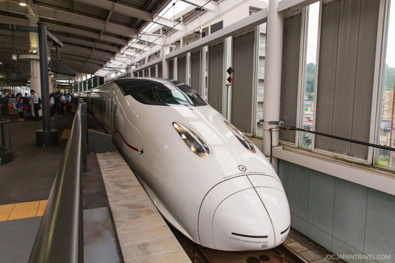 A sleek, white Japanese bullet train is stopped at a station platform under a covered roof, where passengers with Japan train tickets wait nearby and urban scenery is visible through large windows.