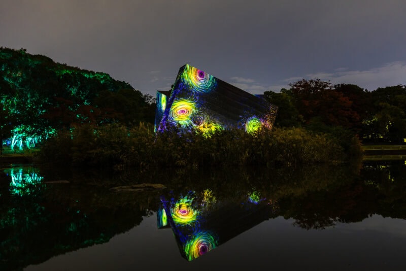 A modern building by a pond at teamLab Botanical Garden Osaka is illuminated at night with swirling, colorful light projections in yellow, green, and purple, their reflections shimmering on the calm water. Trees and bushes surround the enchanting scene.
