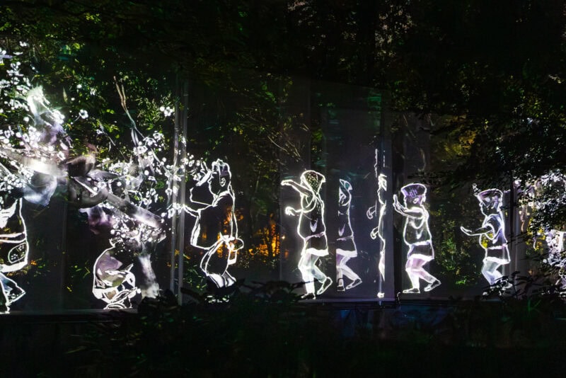 Glowing, animated white line drawings of children appear to walk and play in a dark outdoor setting at teamLab Botanical Garden Osaka, surrounded by trees and illuminated foliage, creating a whimsical and magical atmosphere.