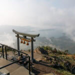 A traditional Japanese torii gate stands on a mountainside, overlooking the misty Amakusa Islands—a must-visit spot offering scenic views of hills, tranquil waters, and distant islands beneath a cloudy sky.