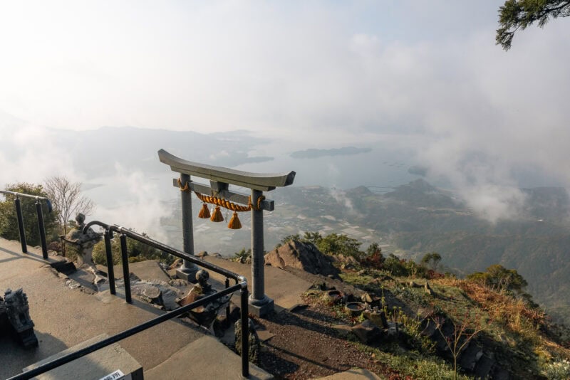A traditional Japanese torii gate stands on a mountainside, overlooking the misty Amakusa Islands—a must-visit spot offering scenic views of hills, tranquil waters, and distant islands beneath a cloudy sky.