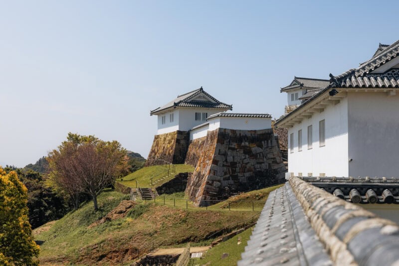 Traditional Japanese castle buildings with white walls and tiled roofs sit atop tall stone foundations, surrounded by grassy hills and trees under a clear blue sky—one of the must-visit spots when exploring things to do in the Amakusa Islands.