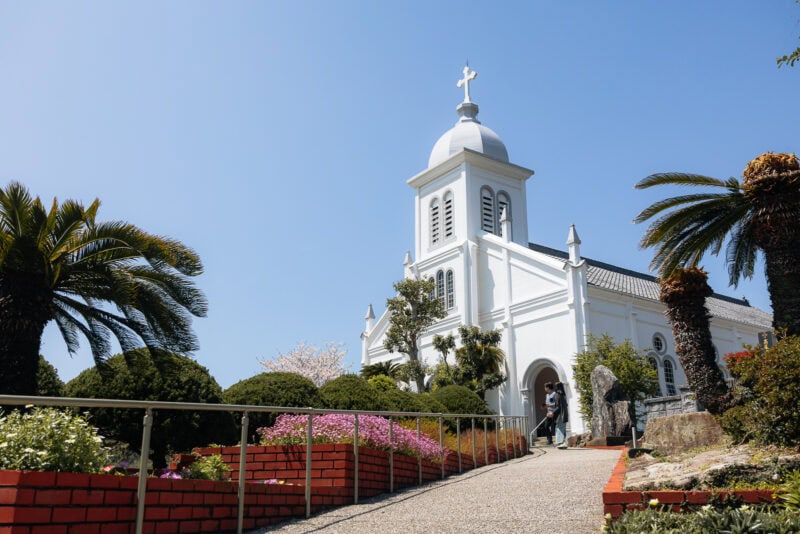 A white church with a cross on its steeple stands under a clear blue sky, surrounded by palm trees and flowering bushes—a must-visit spot among the things to do in the scenic Amakusa Islands. A red brick-bordered path leads to its welcoming entrance.