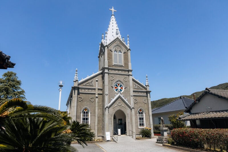 A gray stone church with a tall steeple, white trim, and stained glass windows stands under a clear blue sky—one of the must-visit spots on the Amakusa Islands, surrounded by lush greenery and traditional houses.
