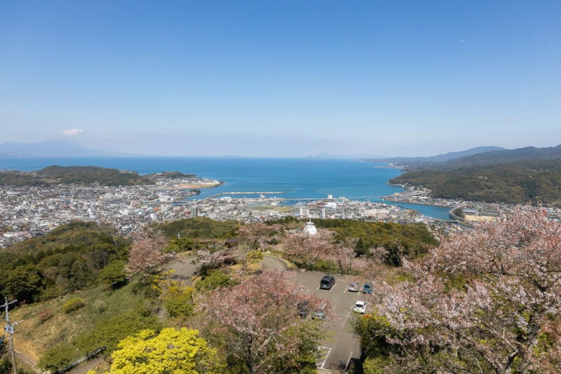 A panoramic view of a coastal city with a harbor in the Amakusa Islands, surrounded by green hills and cherry blossom trees; cars are parked in a small lot, with must-visit spots and the blue ocean stretching to the horizon under a clear sky.
