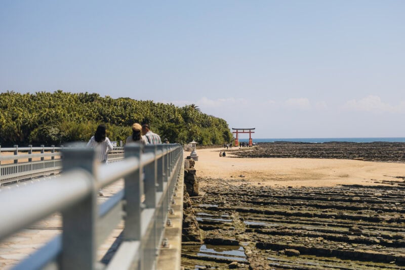 People walk on a bridge toward a sandy beach and a red torii gate on Aoshima Island in Miyazaki, with lush greenery left and rocky shoreline right under a clear blue sky, capturing the beauty of this tropical island destination.