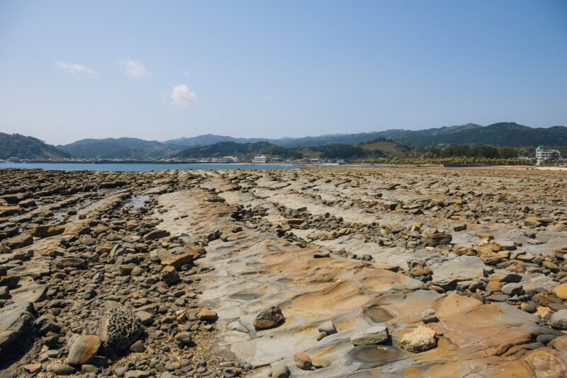 A rocky coastal landscape with flat, layered rock formations in the foreground, reminiscent of Miyazaki’s Aoshima Island, with distant mountains and buildings under a clear blue sky.