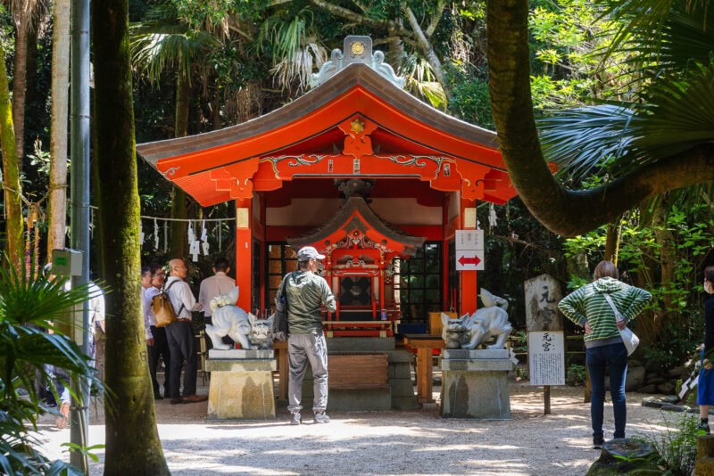 People stand in front of a small red Shinto shrine on Aoshima Island, surrounded by lush greenery and stone guardian statues, as sunlight filters through the trees on this tropical island.