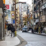 Azabu Juban - Tokyo’s Timeless Neighborhood A busy city street in Azabu Juban, Japan, with people walking, cycling, and shopping. Small shops, signs, and power lines line the narrow road as buildings rise in the background. A car drives down the center of the street.