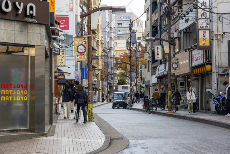 A busy city street in Azabu Juban, Japan, with people walking, cycling, and shopping. Small shops, signs, and power lines line the narrow road as buildings rise in the background. A car drives down the center of the street.