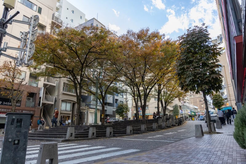 A city street in Azabu Juban with autumn trees lining a pedestrian area. People walk along the sidewalk and crosswalk; cars are parked nearby. Modern buildings and a mostly clear sky with some clouds are in the background.