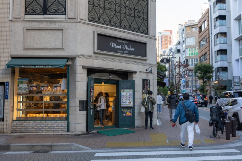 A bakery with a green awning and large window displays bread in Azabu Juban. People enter and exit the shop on this urban street corner, where pedestrians and bikes are nearby.