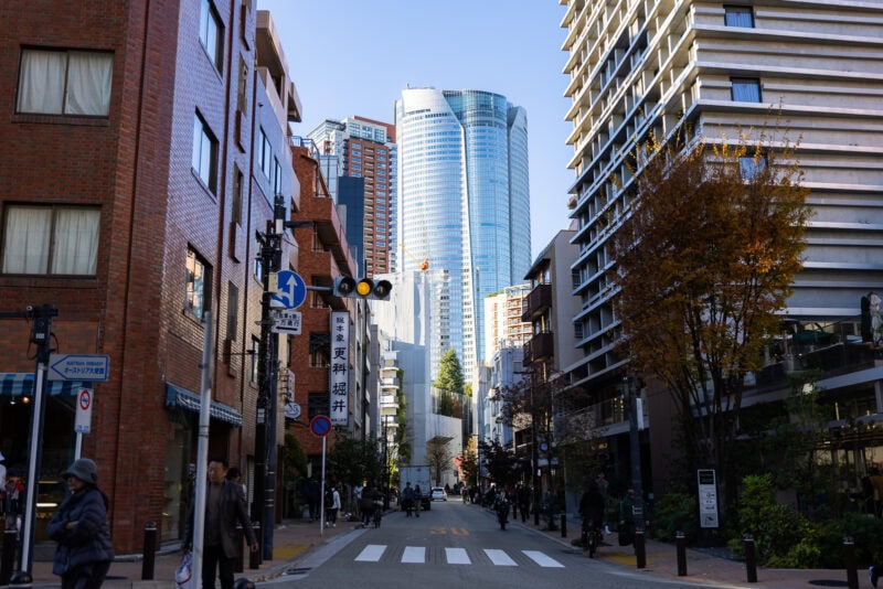 A city street in Azabu Juban, Tokyo, with people walking, tall modern and older brick buildings on both sides, and a large glass skyscraper in the background under a clear sky.
