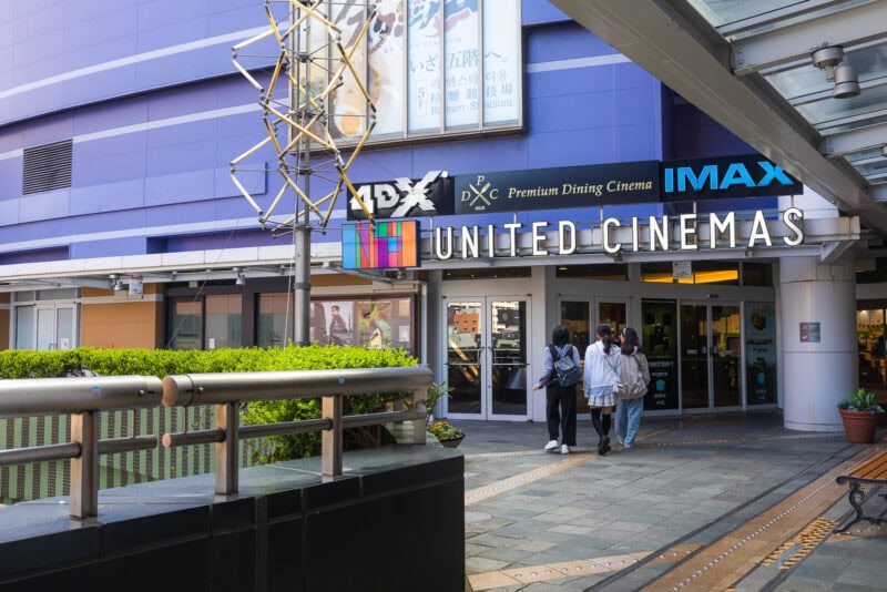 People walk toward the entrance of United Cinemas at Canal City Hakata, a Fukuoka entertainment complex, featuring signage for IMAX, 4DX, and Premium Dining Cinema outside a modern building with purple walls and nearby greenery.