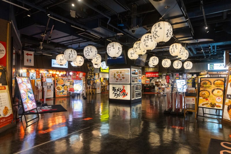 A brightly lit Japanese food court in Canal City Hakata, Fukuoka’s renowned entertainment complex, with hanging white paper lanterns featuring black kanji, illuminated menu boards, and displays of food at various restaurant stalls on a shiny floor.