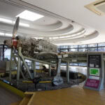 A weathered, partially restored World War II airplane is displayed on supports inside the Chiran Peace Museum, with large windows and informational signs nearby. A person stands to the left, observing this piece of Japan wartime history.