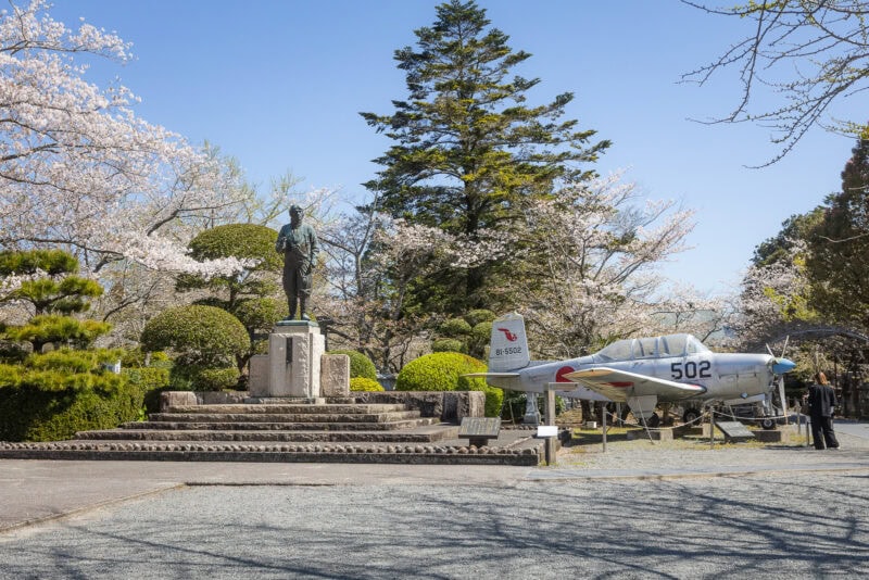 A statue on a stone pedestal stands beside a vintage military aircraft at the Chiran Peace Museum, surrounded by cherry blossom trees in full bloom on a clear, sunny day—a serene tribute to Japan wartime history.