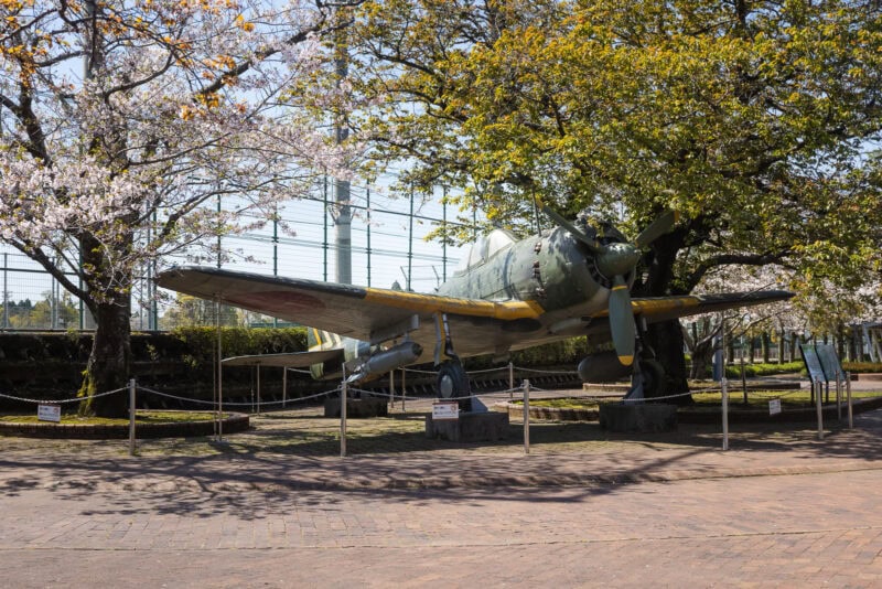 A vintage military aircraft on outdoor display at the Chiran Peace Museum, surrounded by blooming cherry blossom trees and greenery, with informational signs nearby and a brick-paved area in the foreground—evoking reflections on Japan wartime history.