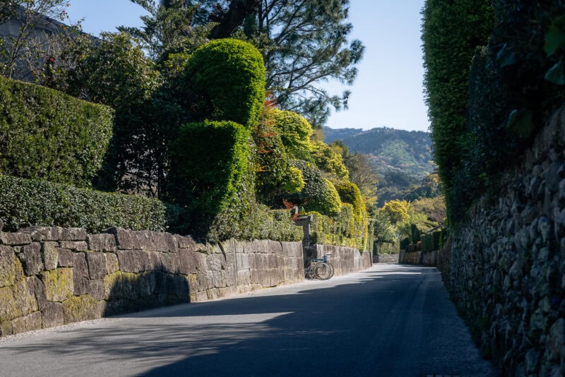 A quiet, sunlit street in the Chiran Samurai District, Japan, bordered by tall hedges and stone walls, with a bicycle parked on the left. Trees and green hills rise in the background under a clear blue sky, evoking echoes of the Samurai Era.