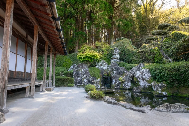 A traditional Japanese rock garden from the Samurai Era, with raked gravel, moss-covered rocks, a small pond, lush greenery, and a wooden building on the left, set against tall trees—evoking the charm of Japan's Chiran Samurai District.