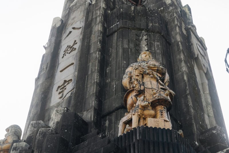 A stone statue of a warrior in armor stands before the Peace Tower at Heiwadai Park, Japan. The monument, with large vertical Japanese kanji inscriptions, features intricate details and a strikingly dark, aged appearance.