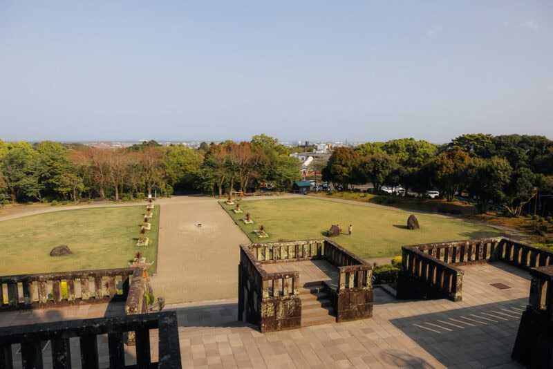 View from a stone terrace overlooking a large, grassy courtyard with scattered rocks and small shrubs at Heiwadai Park in Japan, surrounded by trees and with distant buildings visible beneath a clear sky.