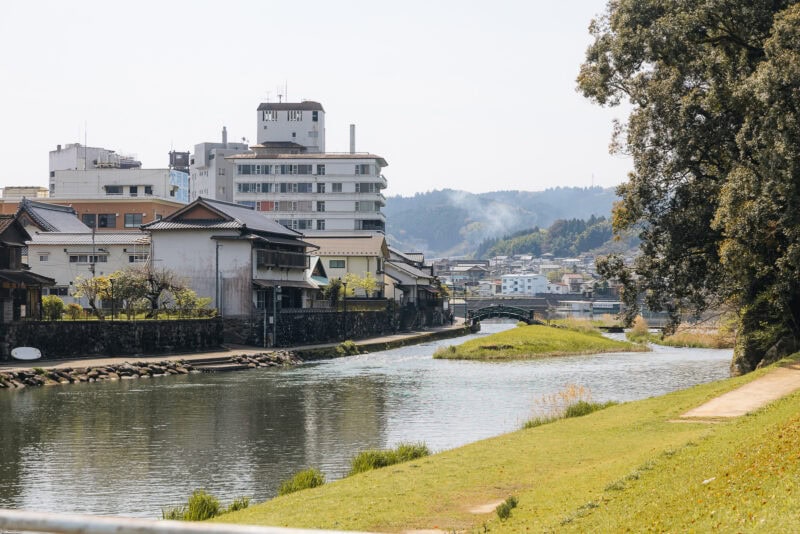 A calm river flows through Hita, known as Little Kyoto in Oita, lined with traditional and modern buildings. A small bridge arches in the distance, with hills beyond and grass and trees bordering the water under a clear sky.