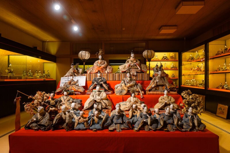 A display of traditional Japanese Hina dolls arranged on red steps, featuring elaborately dressed figures in a museum-like setting in Hita, Oita—Little Kyoto—with additional dolls showcased in illuminated cases along the walls.