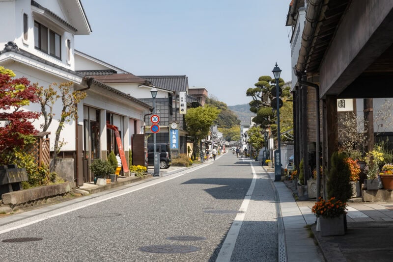 A quiet street in Hita, Oita&mdash;nicknamed Little Kyoto&mdash;features white buildings, potted plants, street signs, and lampposts on a sunny day; greenery and hills form a picturesque backdrop.