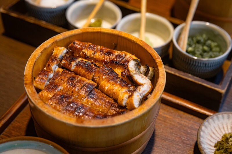 Grilled eel fillets arranged on rice in a wooden bowl, served in Hita—Oita's Little Kyoto—with various small bowls of condiments and accompaniments on a wooden tray.