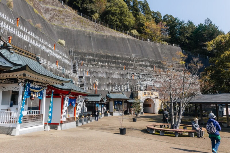 A courtyard at a Japanese temple complex in Hita, Oita—known as Little Kyoto—with statues arranged in rows on a steep hillside, traditional temple buildings, trees, and several people walking or standing around the area.