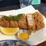 What to Eat in Kagoshima: 7 Must-Try Foods A plate of sliced, crispy breaded pork cutlet (tonkatsu)—a Kagoshima food classic—is served with a lemon wedge, a dollop of mustard in a foil cup, and a garnish of green leaves on a white dish; truly one of the must-try foods here.