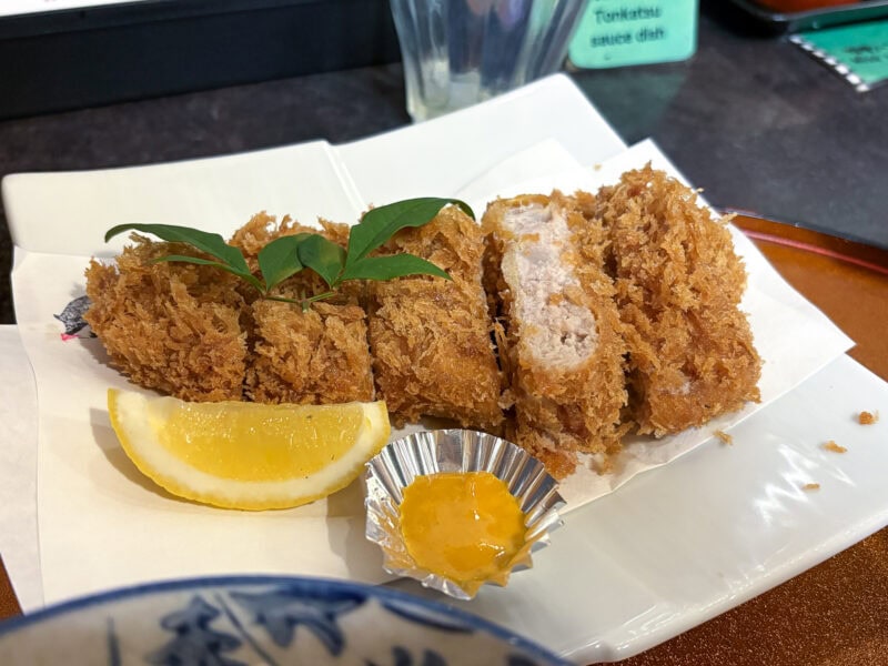 A plate of sliced, crispy breaded pork cutlet (tonkatsu)&mdash;a Kagoshima food classic&mdash;is served with a lemon wedge, a dollop of mustard in a foil cup, and a garnish of green leaves on a white dish; truly one of the must-try foods here.