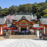 Kirishima Shrine: Kagoshima's Sacred Shrine Traditional Japanese Sacred Shrine with ornate red, white, and gold detailing, surrounded by trees and people walking nearby under a clear blue sky. Cherry blossoms bloom in the background at the historic Kirishima Shrine in Kagoshima.