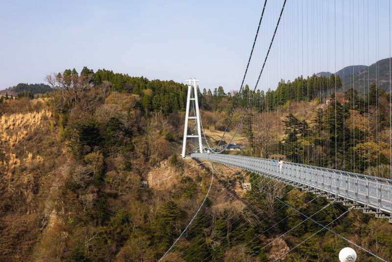 The Kokonoe Yume Suspension Bridge, one of Kyushu's longest bridges, stretches across a deep forested valley, supported by tall white towers, with green trees and hills in the background under a clear sky.