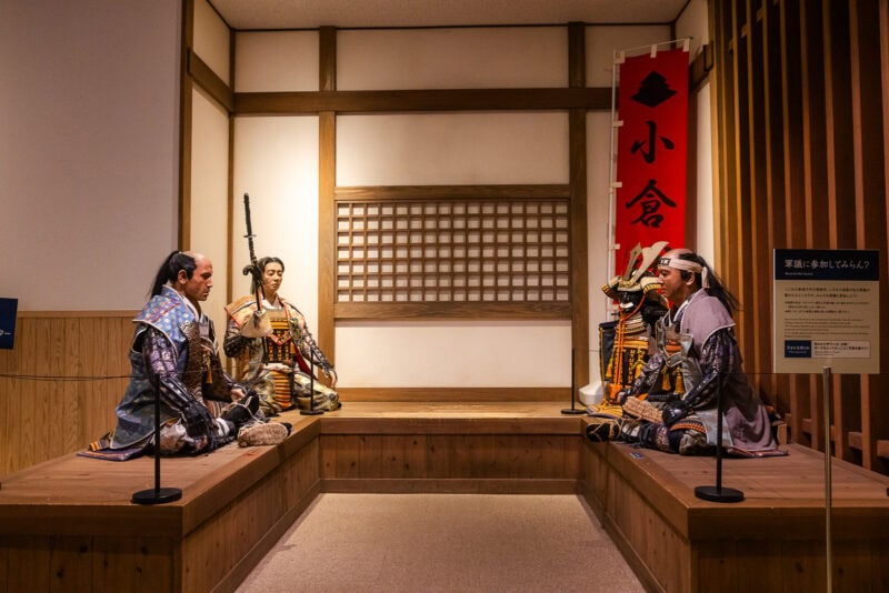 Three life-sized mannequins dressed as samurai sit in a traditional Japanese room at Kokura Castle, engaging in conversation. The setting features wooden walls, tatami mats, and a red banner—immersing visitors in Kitakyushu's samurai heritage.