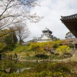 Kokura Castle: A Symbol of Kitakyushu's Samurai Heritage Kokura Castle, symbolizing Kitakyushu's Samurai Heritage, stands on a hill surrounded by autumn trees, a pond, and a wooden building in the foreground beneath a partly cloudy sky.