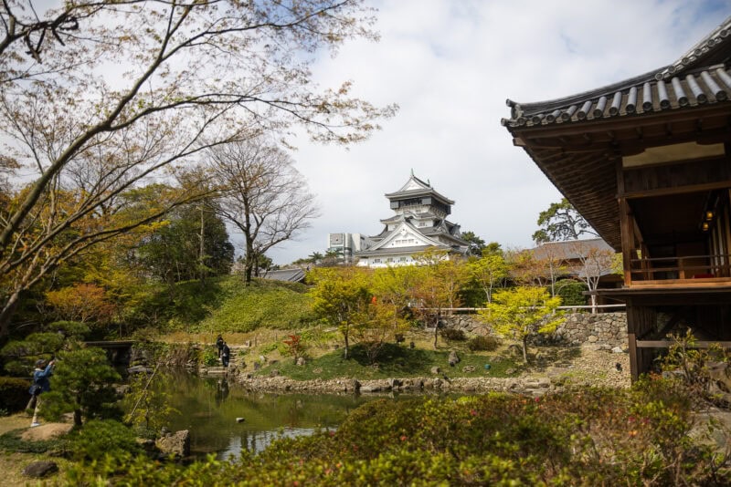 Kokura Castle, symbolizing Kitakyushu's Samurai Heritage, stands on a hill surrounded by autumn trees, a pond, and a wooden building in the foreground beneath a partly cloudy sky.