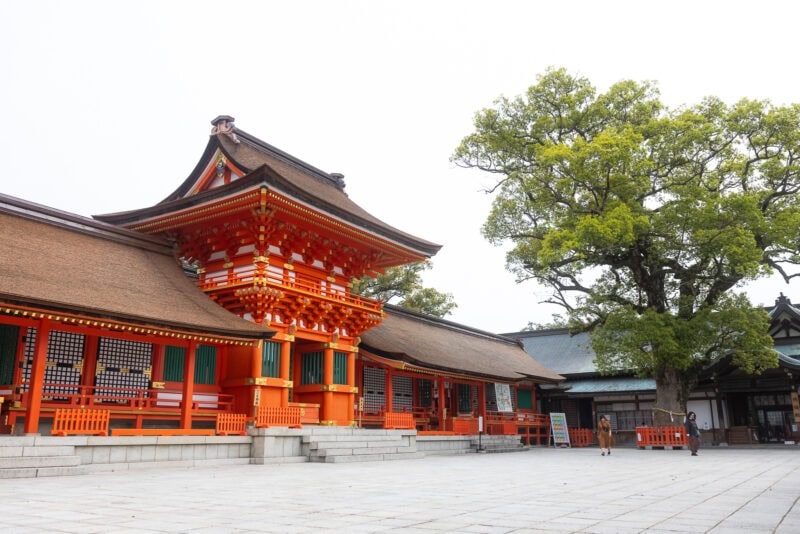 A traditional Japanese shrine on the Kunisaki Peninsula in Oita, with bright orange and white wooden structures, sloped roofs, and a large green tree on a spacious stone courtyard under a clear sky in this sacred region.