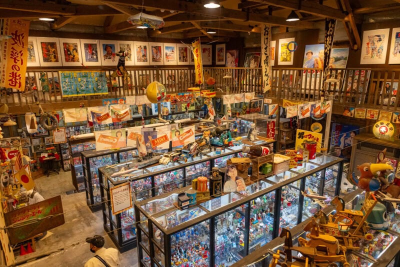 A colorful, nostalgic Japanese toy store in Oita’s Kunisaki Peninsula, filled with vintage toys, posters, and memorabilia. Glass display cases and shelves are packed with toys under warm lighting in this Sacred Region-inspired shop.