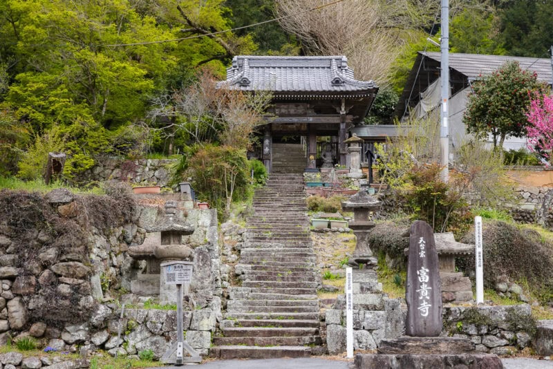 Stone steps lead up to a traditional wooden Japanese temple gate on the tranquil Kunisaki Peninsula in Oita, surrounded by greenery, stone lanterns, and signs with Japanese writing in this sacred region.