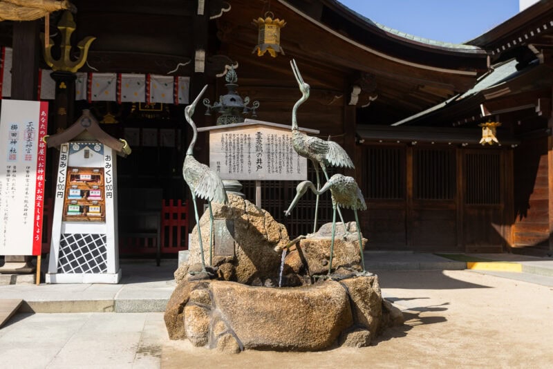 A bronze crane statue fountain stands on a rock base in front of a wooden Japanese shrine building at Kushida Shrine, the oldest shrine in Fukuoka, beneath a sign with Japanese text on a sunny day.