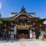 Kushida Shrine: Fukuoka's Oldest Shrine A traditional Japanese shrine with ornate wooden architecture and a large rope decoration above the entrance. Several people are standing or walking near Kushida Shrine in Fukuoka on a sunny day with a blue sky.
