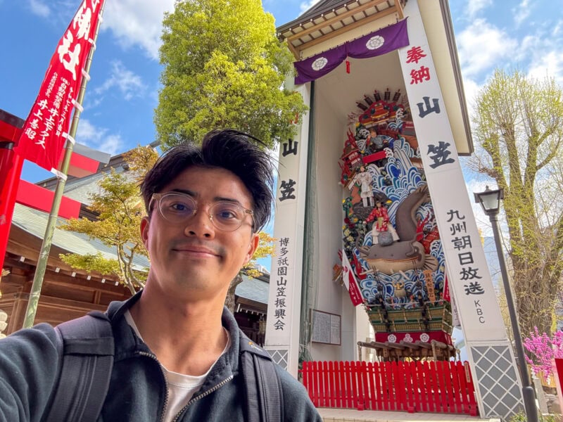 A person takes a selfie in front of a tall, ornate Japanese festival float display at Fukuoka’s Kushida Shrine—the oldest shrine in the city—with red banners, trees, and a bright blue sky in the background.