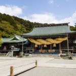 Miyajidake Shrine: The Path of Light A traditional Japanese Miyajidake Shrine with a large, thick shimenawa above the entrance, green-tiled roofs, stone lanterns, and trees under a partly cloudy sky—famed for its Path of Light.
