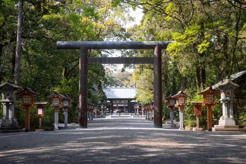 A large traditional Japanese torii gate stands at the entrance of a shaded path lined with stone lanterns, leading to Miyazaki Shrine, one of the oldest shrines in Miyazaki, surrounded by lush green trees.