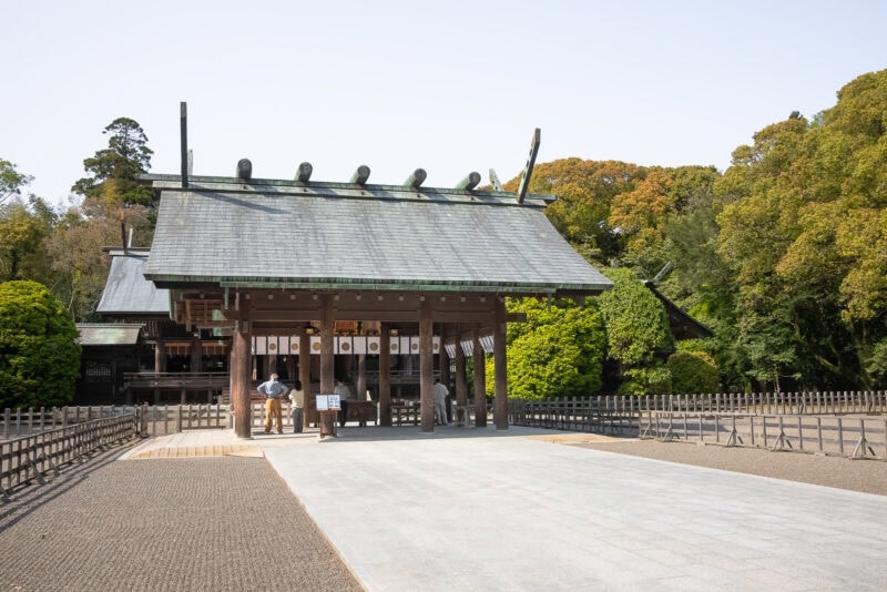 A traditional Japanese shrine, Miyazaki Shrine, features a large wooden gate, sloped green roof, and decorative beams, surrounded by trees and a gravel courtyard under a clear sky in Miyazaki.