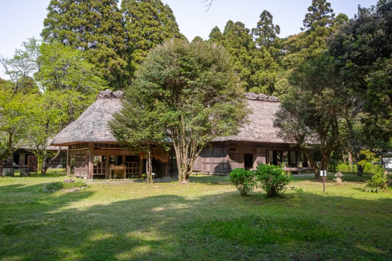 A traditional Japanese farmhouse with a thatched roof is surrounded by lush trees and greenery on a sunny day, evoking the serene landscapes often seen in Miyazaki films. The house sits peacefully among bushes and tall trees in the background.